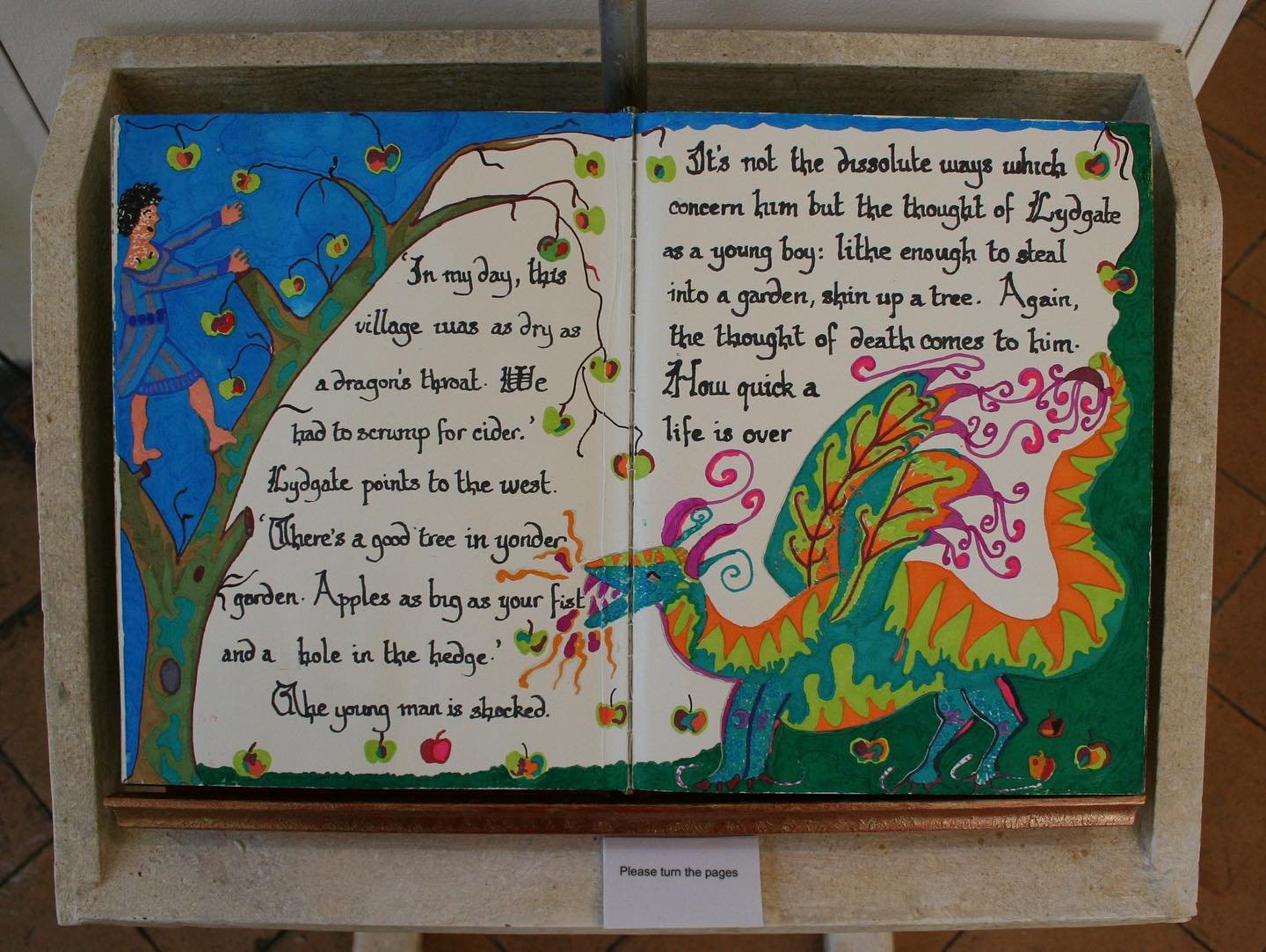 An open hand-illuminated book on a lectern, showing painted figures and calligraphic text — from a Lockarts educative arts and heritage workshop.